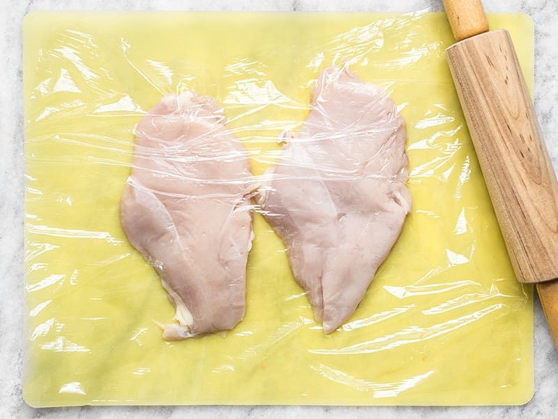 Chicken breast on a cutting board, covered in plastic wrap, with a rolling pin on the side