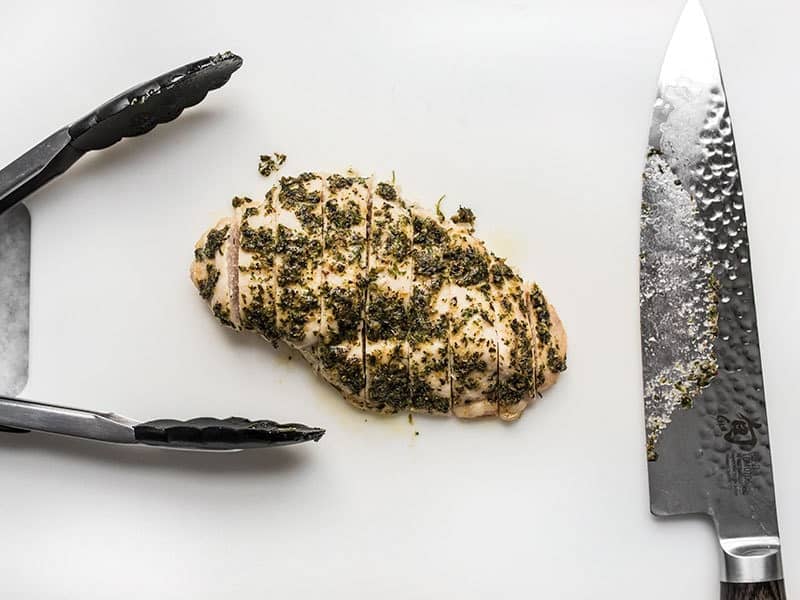 Sliced garlic herb chicken breast on a cutting board with tongs and a knife