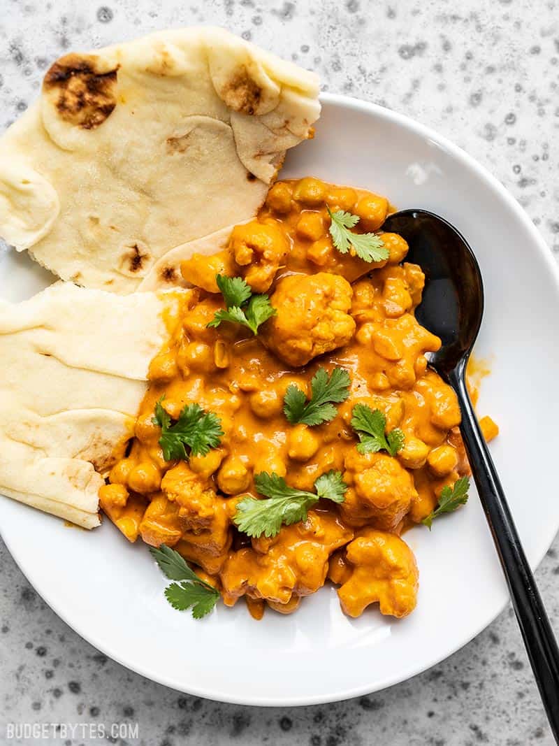 A bowl full of Cauliflower and Chickpea Masala with a black spoon and torn piece of naan bread.