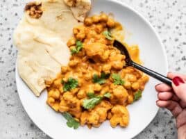 A bowl of creamy Cauliflower and Chickpea Masala being eaten with a spoon and two pieces of naan on the side.