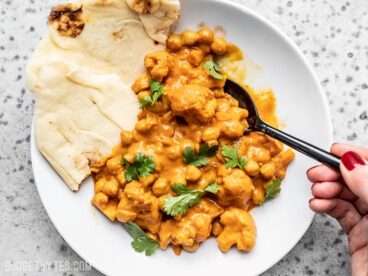 A bowl of creamy Cauliflower and Chickpea Masala being eaten with a spoon and two pieces of naan on the side.