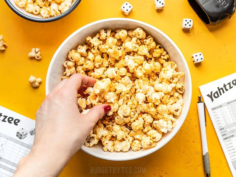 Sriracha Nooch Popcorn is the perfect salty snack with tons of crunch, a spicy buttery coating, and a slightly cheesy flavor thanks to nutritional yeast. Budgetbytes.com A hand grabbing some Sriracha Nooch Popcorn out of a large bowl on a yellow background.