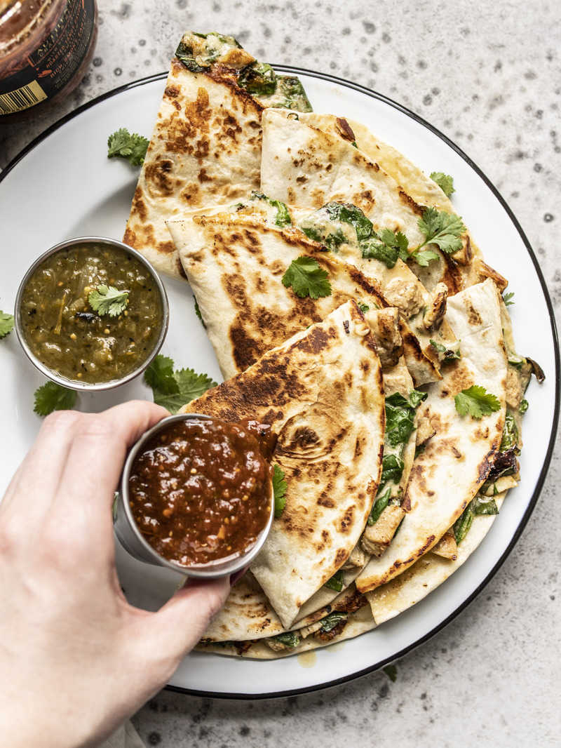 A hand pouring red salsa onto a plate of creamy chicken and spinach quesadillas