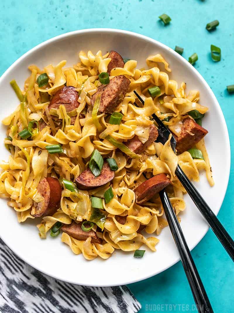 Cajun Cabbage and Noodles is a fun spin on the classic comfort food with a handful of Cajun spices, some smoked sausage, and a splash of hot sauce. Budgetbytes.com A bowl of Cajun Cabbage and Noodles with a black spoon and fork stuck in the side of the bowl. Green onions sprinkled on top.