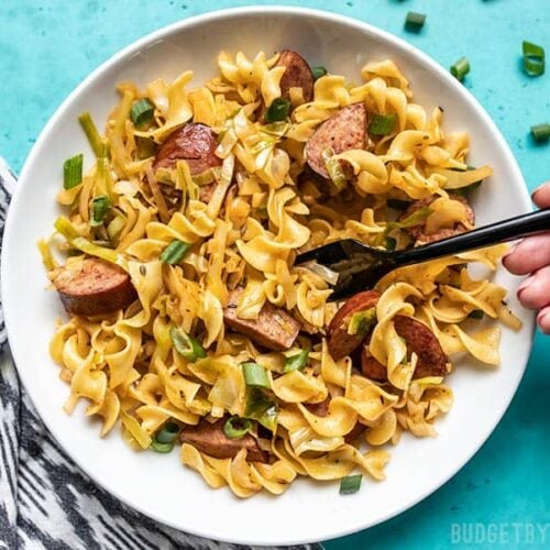 A hand lifting a forkful of Cajun Cabbage and Noodles from a large bowl.