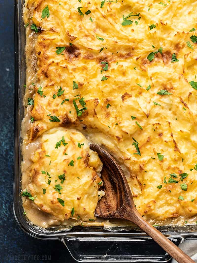 Close up overhead view of Cheesy Cottage Pie being scooped out of the glass casserole dish with a large wooden spoon.