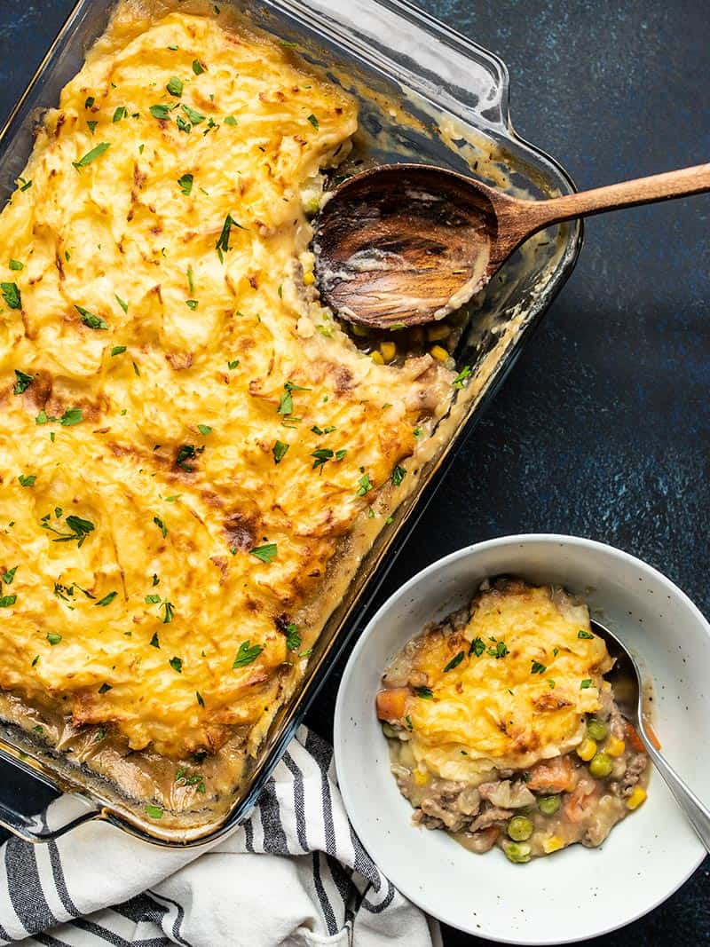 A casserole dish full of Cheesy Cottage Pie with a portion scooped out into a bowl on the side