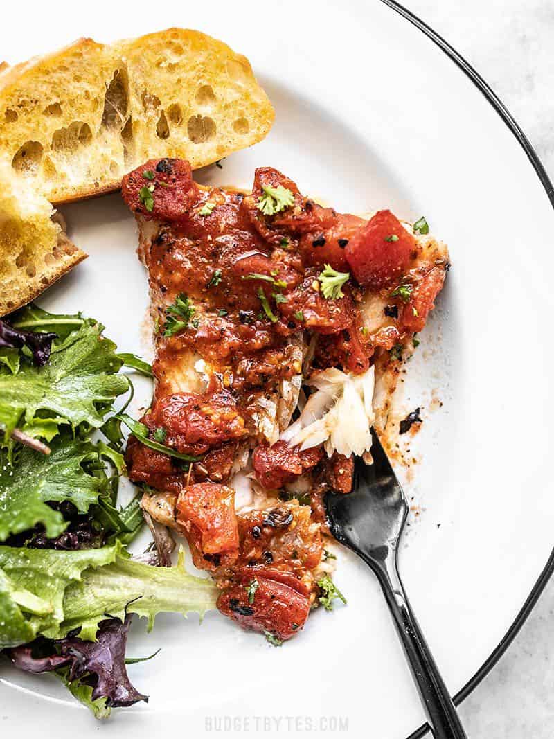 A piece of Easy Oven Baked Fish with Tomatoes on a plate with baguette slices, a green salad, and black fork