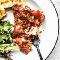 A piece of Easy Oven Baked Fish with Tomatoes on a plate with baguette slices, a green salad, and black fork