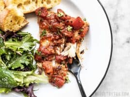 A piece of Easy Oven Baked Fish with Tomatoes on a plate with baguette slices, a green salad, and black fork