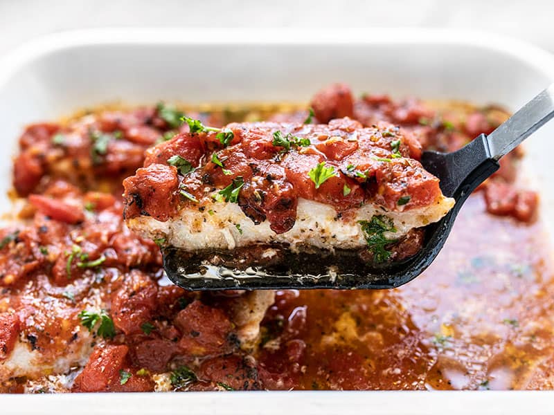 A fillet of baked fish with tomatoes being lifted out of the baking dish with a spatula