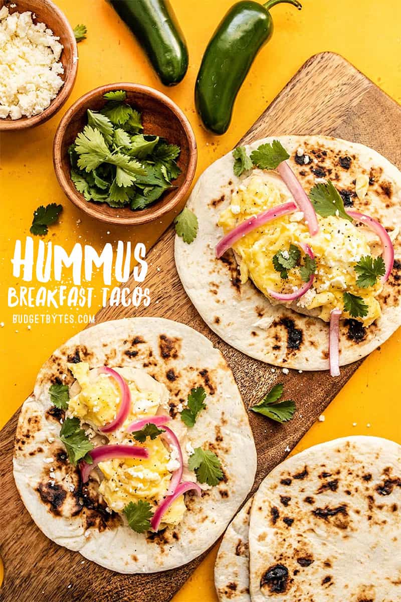 Two Hummus Breakfast Tacos on a wood cutting board with tortillas, cilantro, and feta near by.