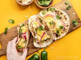 Three Hummus Breakfast Tacos on a wooden cutting board, a hand picking one up