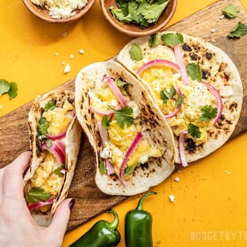 Three Hummus Breakfast Tacos on a wooden cutting board, a hand picking one up