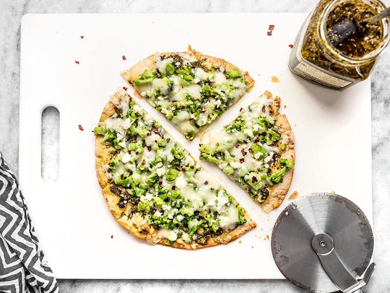 Sliced Quick Fix Broccoli Pesto Pizza on a cutting board with a jar of pesto and pizza wheel on the side.