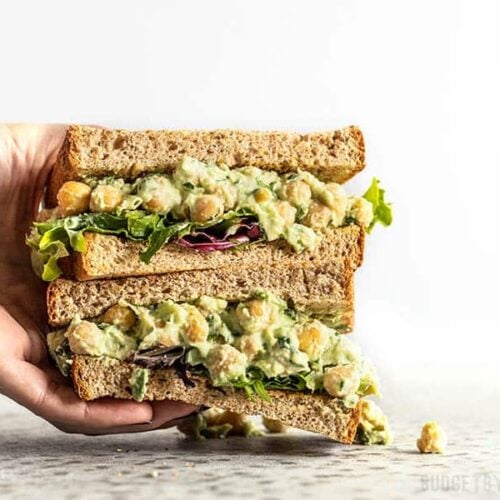 A hand grabbing a stack of two halves of a Scallion Herb Chickpea Salad sandwich on wheat bread.