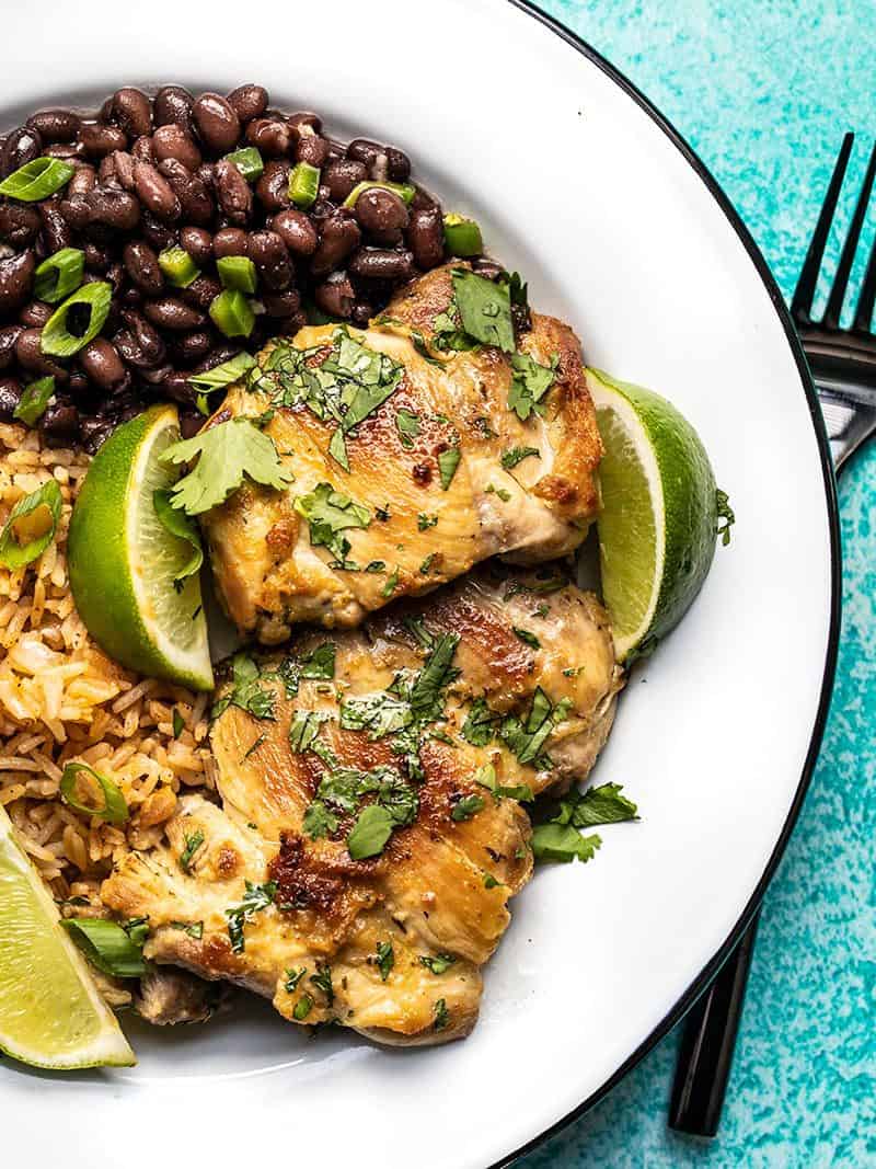 Close up of cilantro lime chicken thighs on a plate with black beans, rice, and lime wedges. 