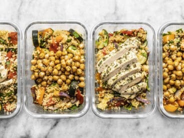 Four glass meal prep containers lined up with roasted vegetable couscous. Two with chicken and two with chickpeas