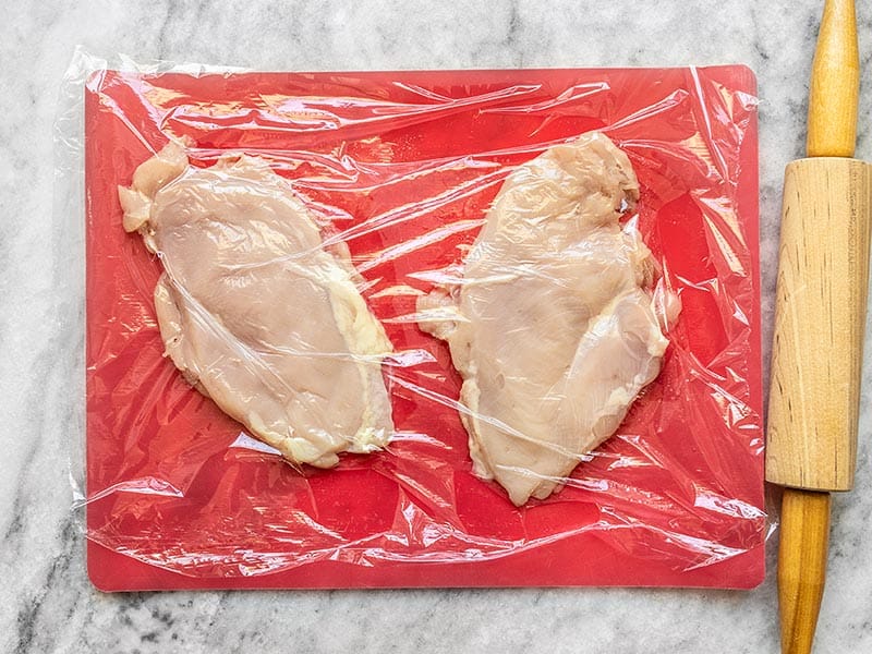 Pounded chicken breasts on a cutting board covered with plastic, rolling pin on the side