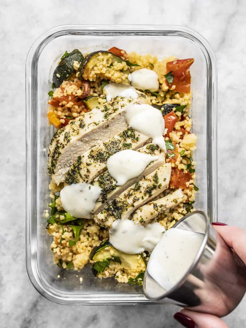 A glass meal prep container with roasted vegetable couscous and sliced chicken, with ranch being drizzled over top.