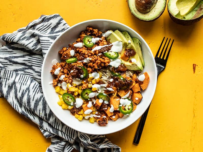 Overhead view of a finished tempeh burrito bowl on a yellow background with a black and white napkin and an avocado on the side.