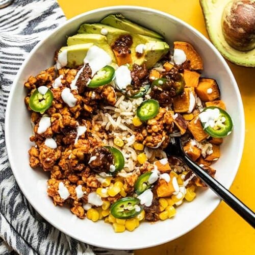 Overhead view of a tempeh burrito bowl with a black fork digging in, an avocado on the side.