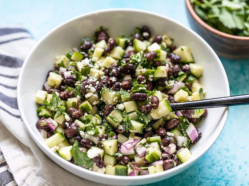 Front view of Cucumber and Black Bean Salad in a white bowl with a black spoon.