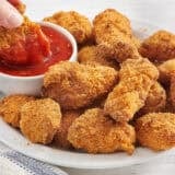 Side view of a plate of homemade chicken nuggets with a hand dipping a nugget into a pot of ketchup.
