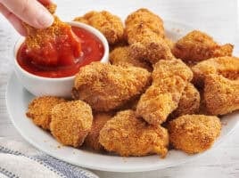 Side view of a plate of homemade chicken nuggets with a hand dipping a nugget into a pot of ketchup.