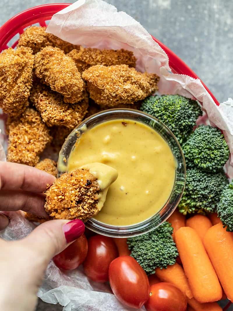 One Homemade Baked Chicken Nugget being dipped into honey mustard, surrounded by more nuggets and vegetables. 