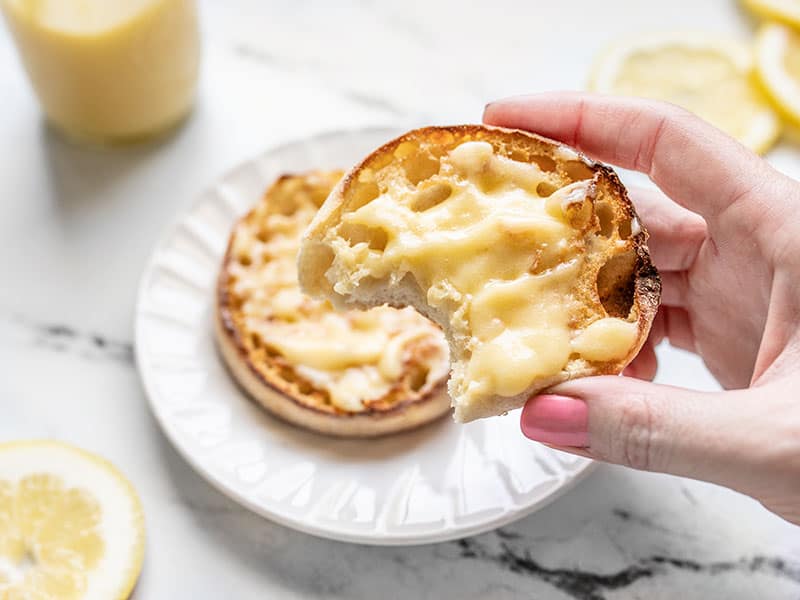 A hand holding an English muffin smeared with homemade lemon curd, a bite is taken out of the muffin.