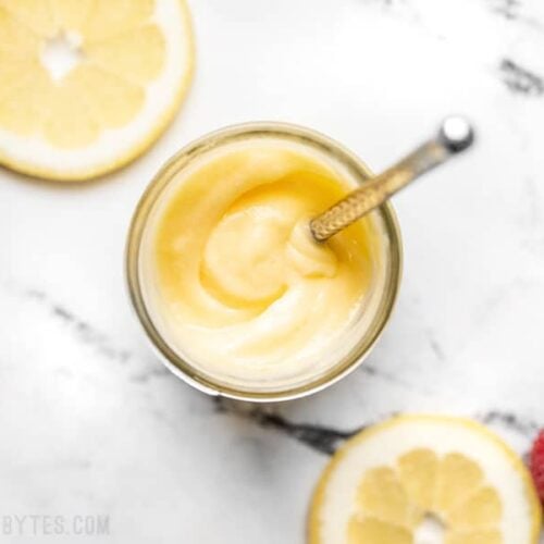 Overhead view of homemade lemon curd in a jar with a small silver butter knife stuck inside, lemon slices on the side.