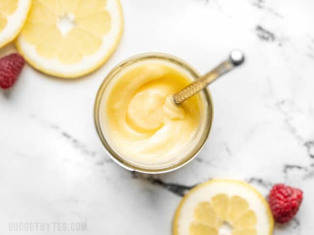 Overhead view of homemade lemon curd in a jar with a small silver butter knife stuck inside, lemon slices on the side.