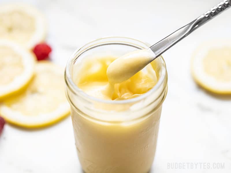 Homemade lemon curd being lifted out of a jar with a small silver butter knife, lemon slices in the background.