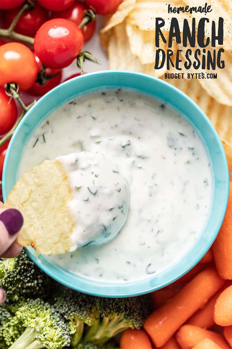 A chip being dipped into a bowl of homemade ranch dressing surrounded by vegetables.