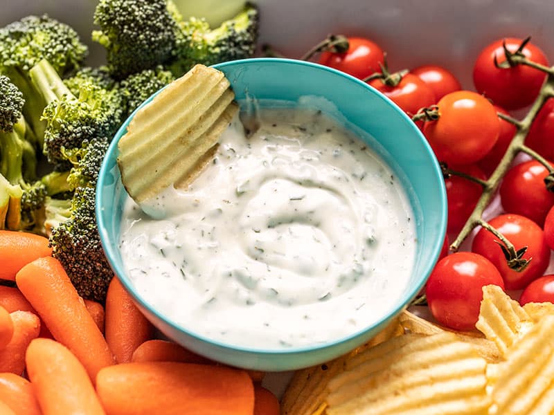 A chip sitting in a bowl of homemade ranch dip surrounded by vegetables and chips