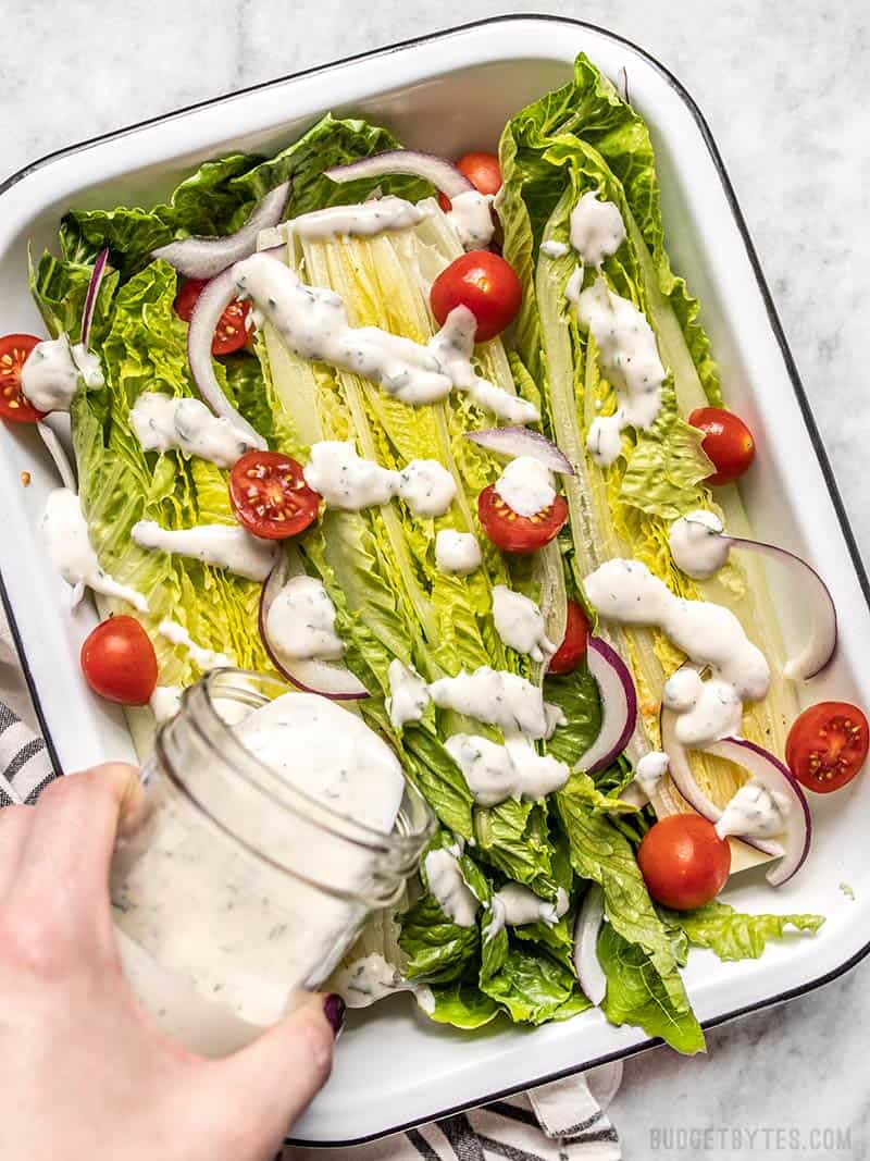 Homemade ranch dressing being drizzled onto a romaine salad with tomatoes and onions.