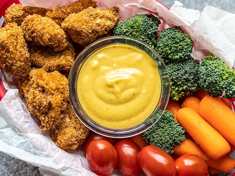 Honey mustard sauce in a dish in the middle of a basket of chicken nuggets and vegetables.