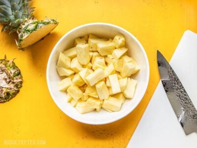 A bowl of fresh pineapple chunks next to a cutting board, chef's knife, and a pineapple top.