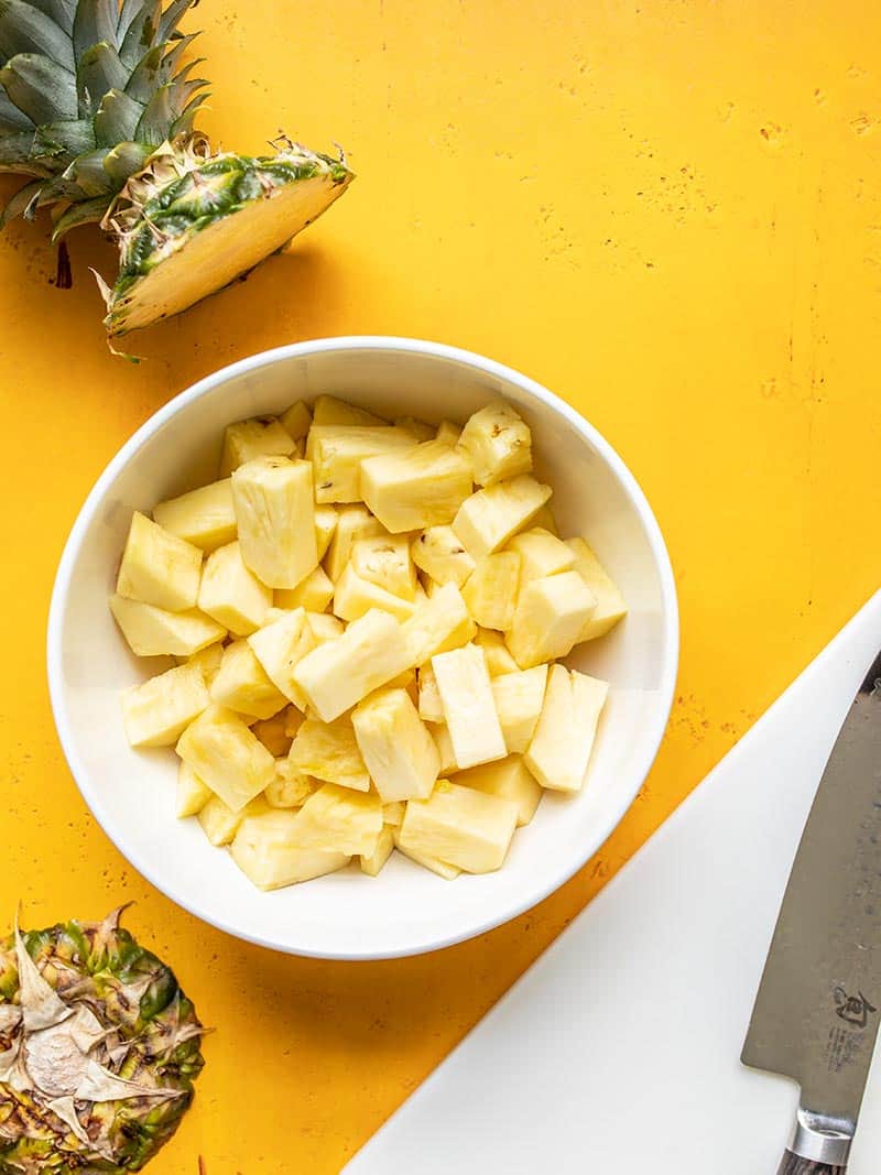 Cut pineapple in a white bowl next to a cutting board and the top and bottom pieces of the fresh pineapple.