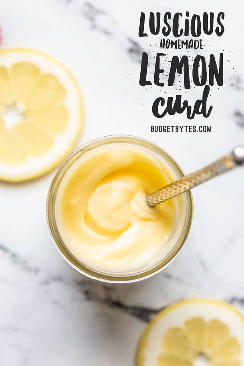 Overhead view of a jar of lemon curd with a small butter knife inside and lemon slices on the sides.