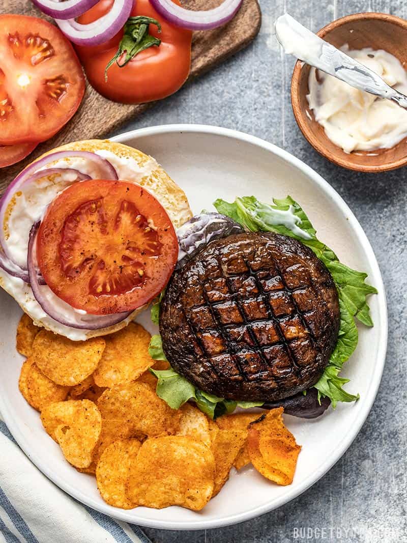 With their deep savory flavor, Marinated Portobello Mushroom Burgers are a fast and easy vegetarian option for summer grilling. Budgetbytes.com An open Marinated Portobello Mushroom Burger on a plate with chips, mayo, sliced tomato, and red onion rings on the side.