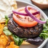 Close up side view of a marinated portobello mushroom burger topped with a slice of tomato and two red onion rings.
