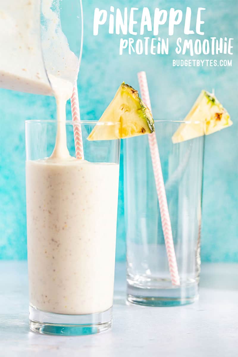 Pineapple Protein Smoothie being poured into a glass with a paper straw and a pineapple wedge on the rim.