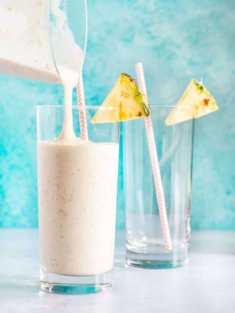 Pineapple Protein Smoothie being poured into a glass with a paper straw and pineapple wedge on the rim.