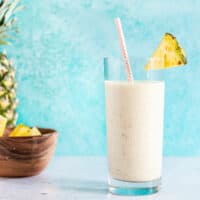 A pineapple protein smoothie in a tall glass with a paper straw, next to a whole pineapple and a small bowl of pineapple chunks