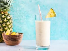 A pineapple protein smoothie in a tall glass with a paper straw, next to a whole pineapple and a small bowl of pineapple chunks