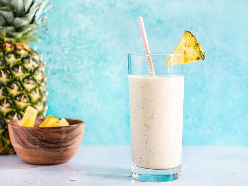 A pineapple protein smoothie in a tall glass with a paper straw, next to a whole pineapple and a small bowl of pineapple chunks