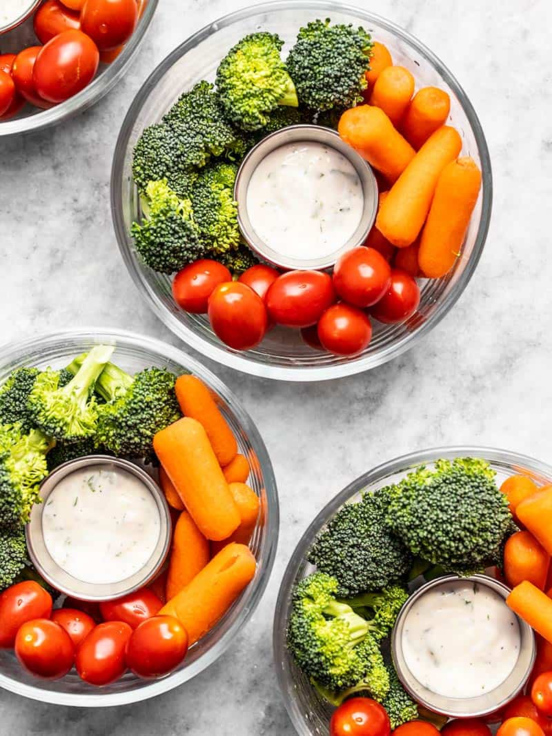 Four glass meal prep bowls made into veggie snack packs with ranch dressing, broccoli, tomatoes, and carrots. 