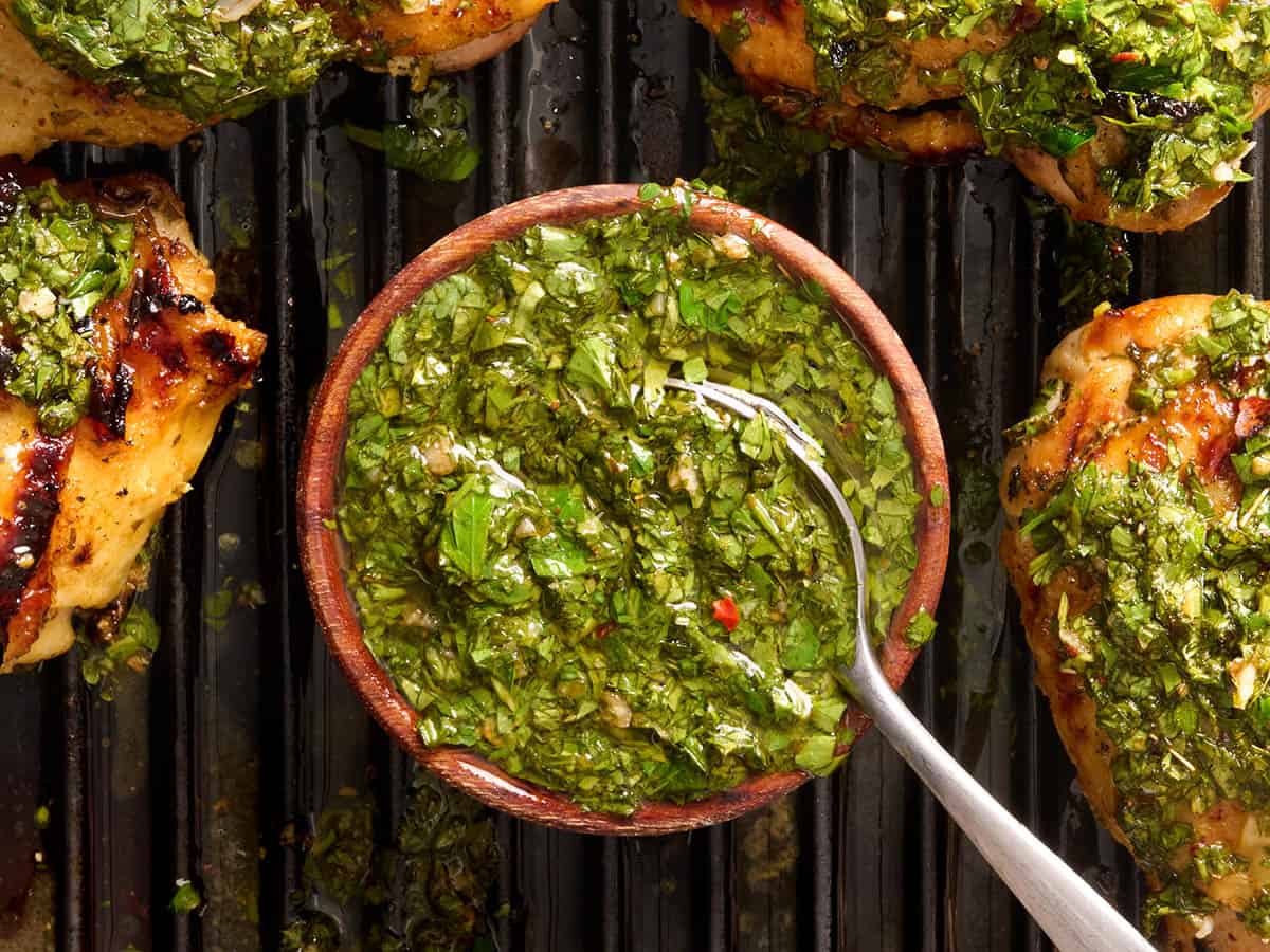 Overhead view of a bowl of chimichurri on a grill with a poon and grilled chicken thighs.